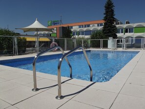 A swimming pool with metal ladders, a covered seating area, and a Holiday Inn Express building in the background. - Holiday Inn Express Strasbourg - Sud by IHG (Geispolsheim)