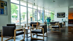 A modern indoor seating area with large windows, wooden chairs, and a teal-colored counter. - Holiday Inn Express Strasbourg - Sud by IHG (Geispolsheim)