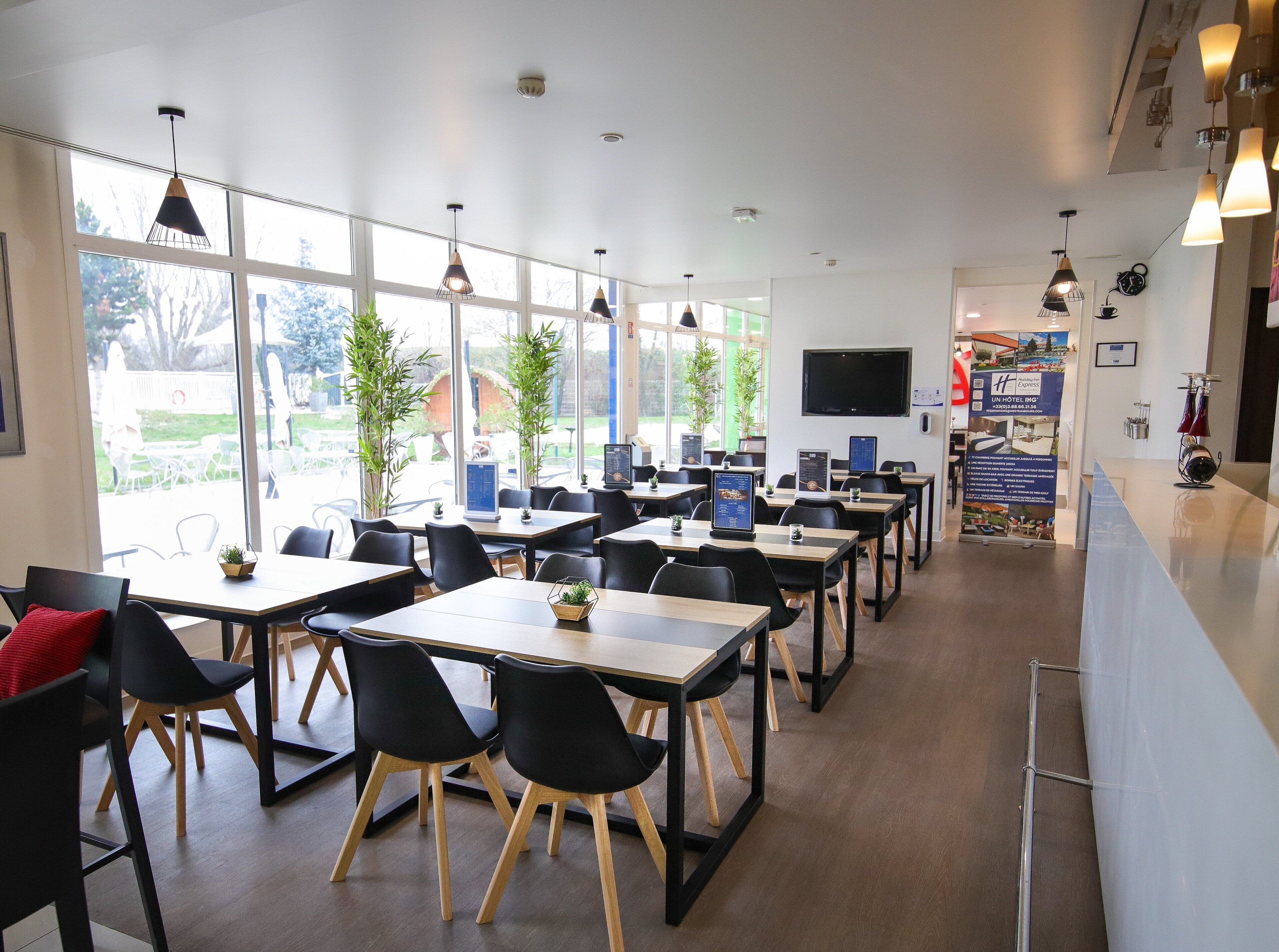 A modern hotel dining area with tables and chairs, large windows, and a reception desk.