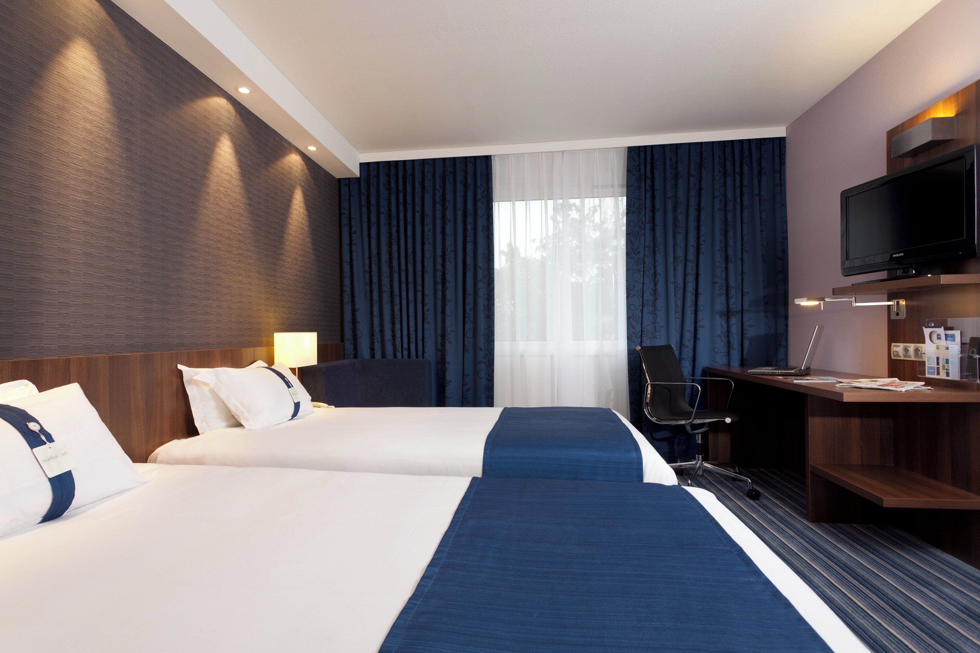 A hotel room with two beds, a desk with a computer, a TV, and a window with curtains.