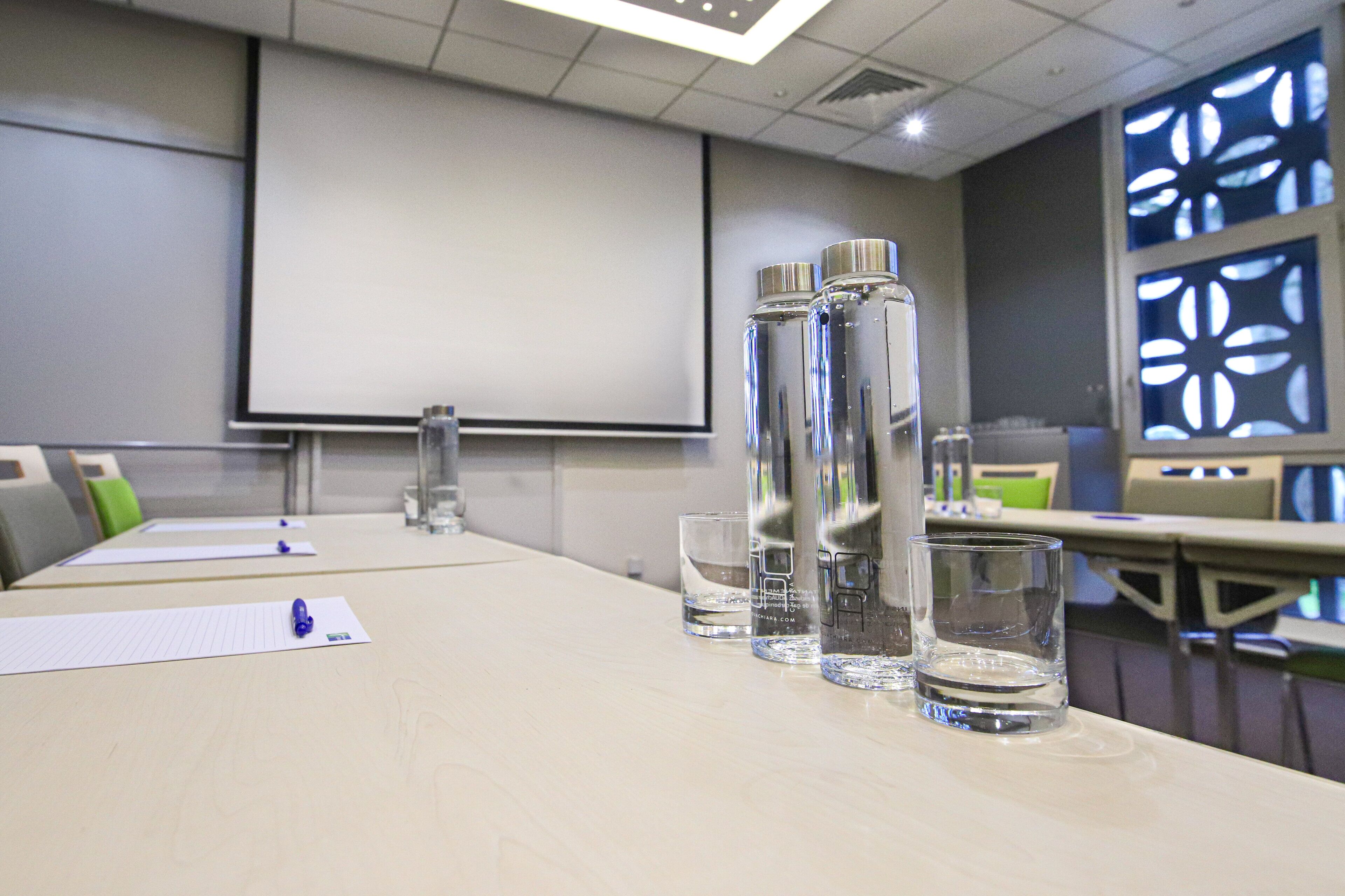 Un salón de conferencias con una larga mesa, botellas de agua, vasos y una pantalla de proyector.