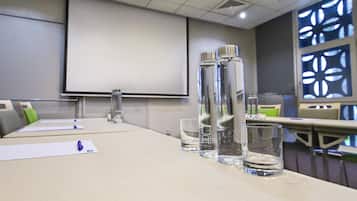 Un salón de conferencias con una larga mesa, botellas de agua, vasos y una pantalla de proyector.