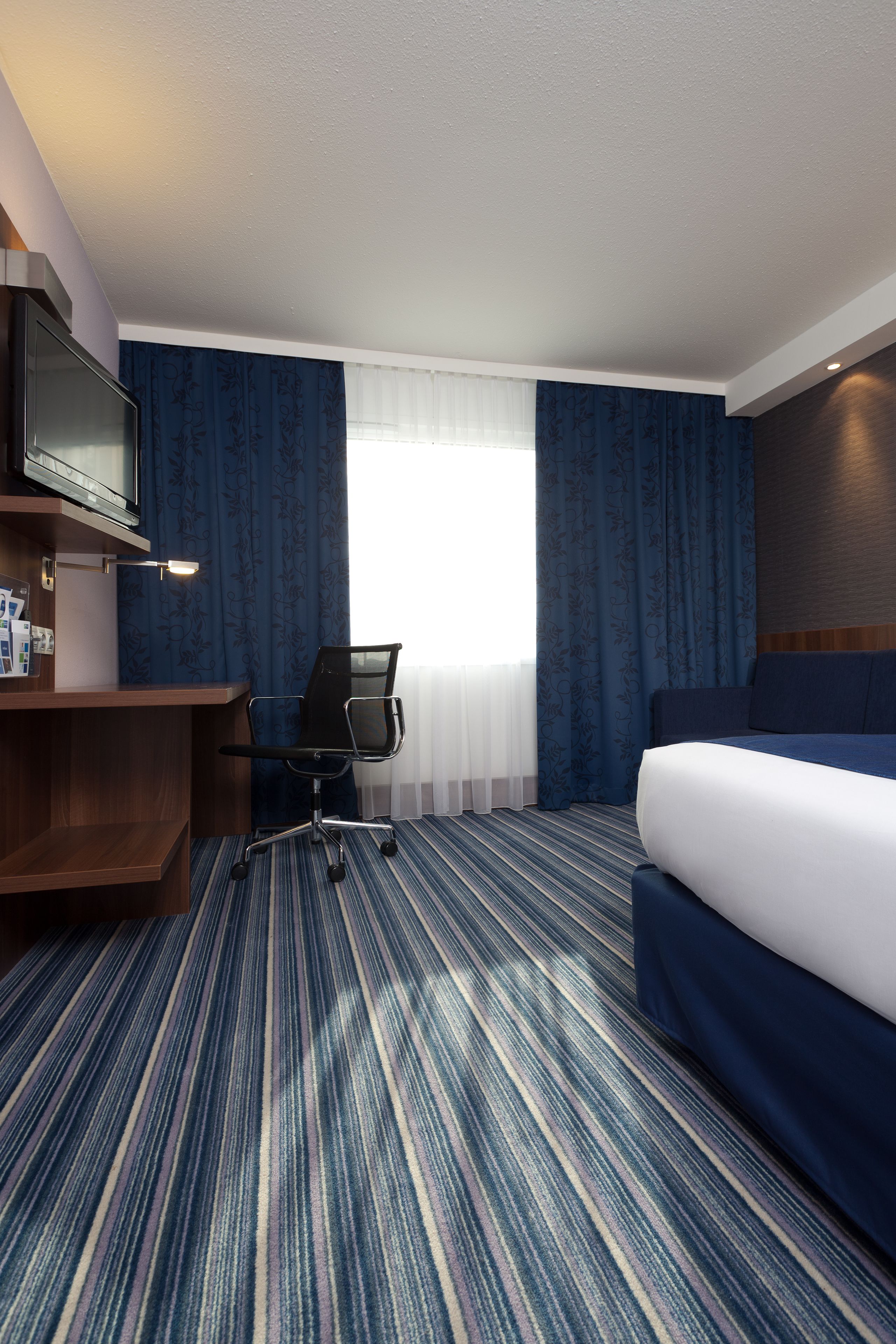 Une chambre d’hôtel comprenant un lit, un bureau avec une chaise, une télévision et une fenêtre avec des rideaux.