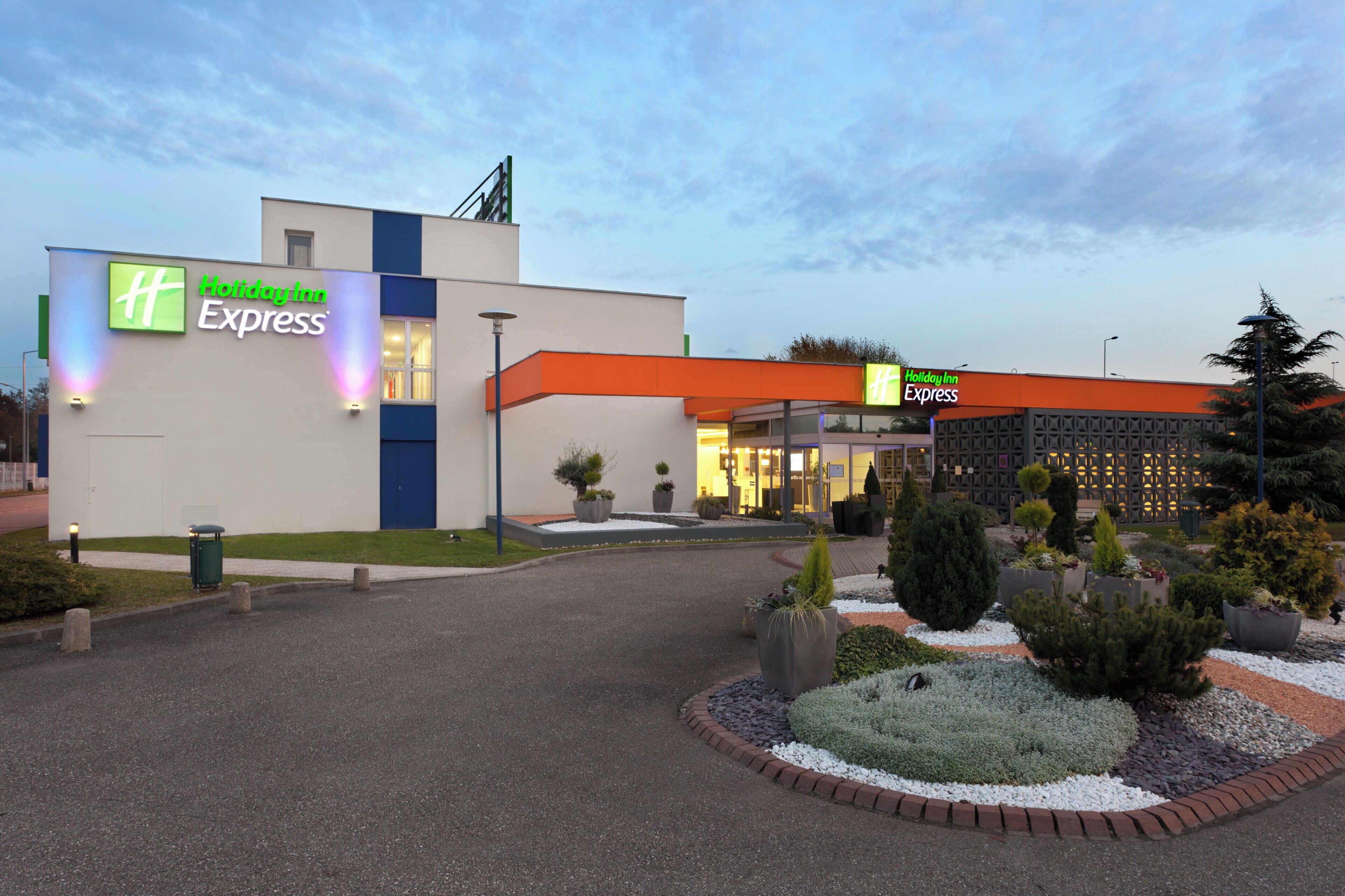 Un hotel Holiday Inn Express con una fachada moderna, un jardín ajardinado y una entrada bien iluminada.