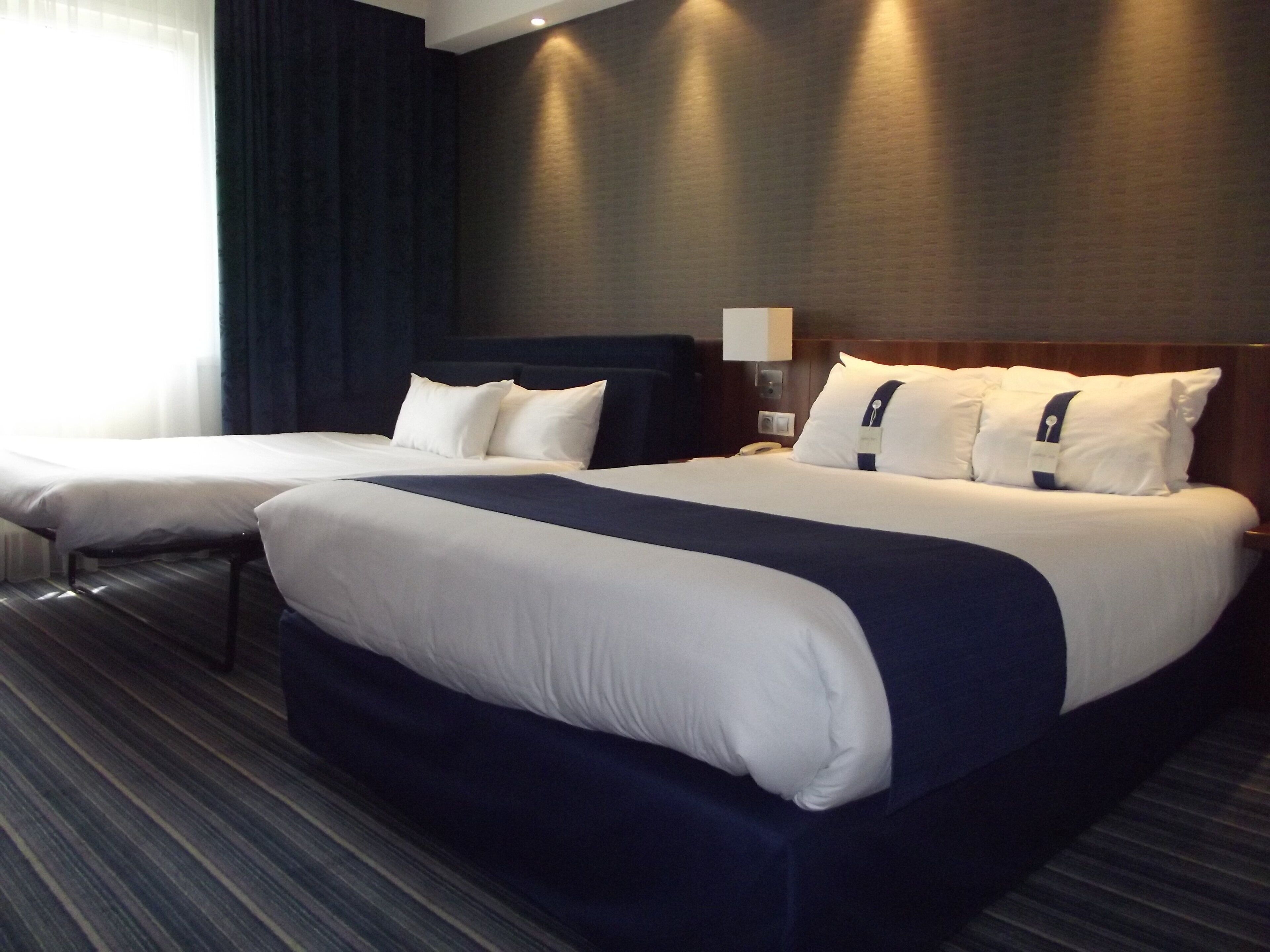 Een hotelkamer met twee bedden, een donker hoofdeinde, een nachtkastje met een lamp en een gestreept tapijt.