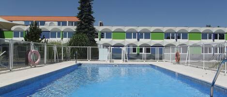 A swimming pool with a clear blue water surface, surrounded by a white fence, and a building with a green and white facade in the background.