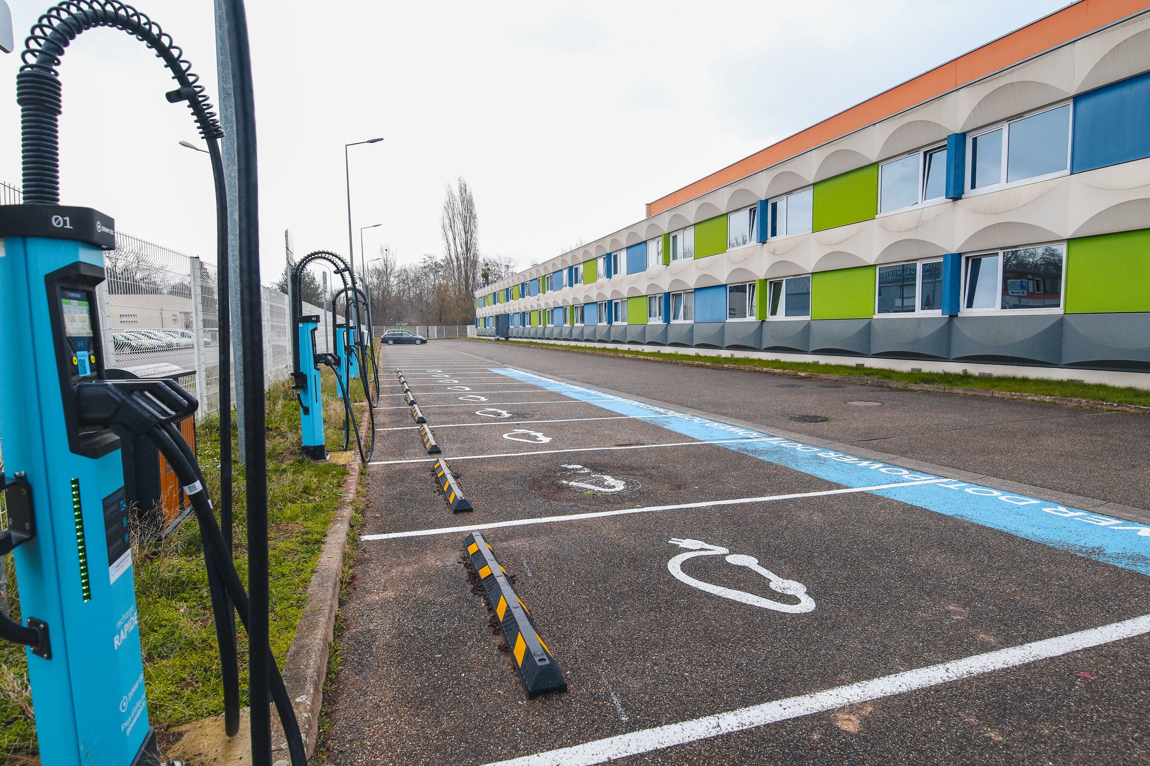 Een parkeerplaats met laadpalen voor elektrische voertuigen, gemarkeerde parkeerplaatsen en een gebouw met een modern ontwerp.