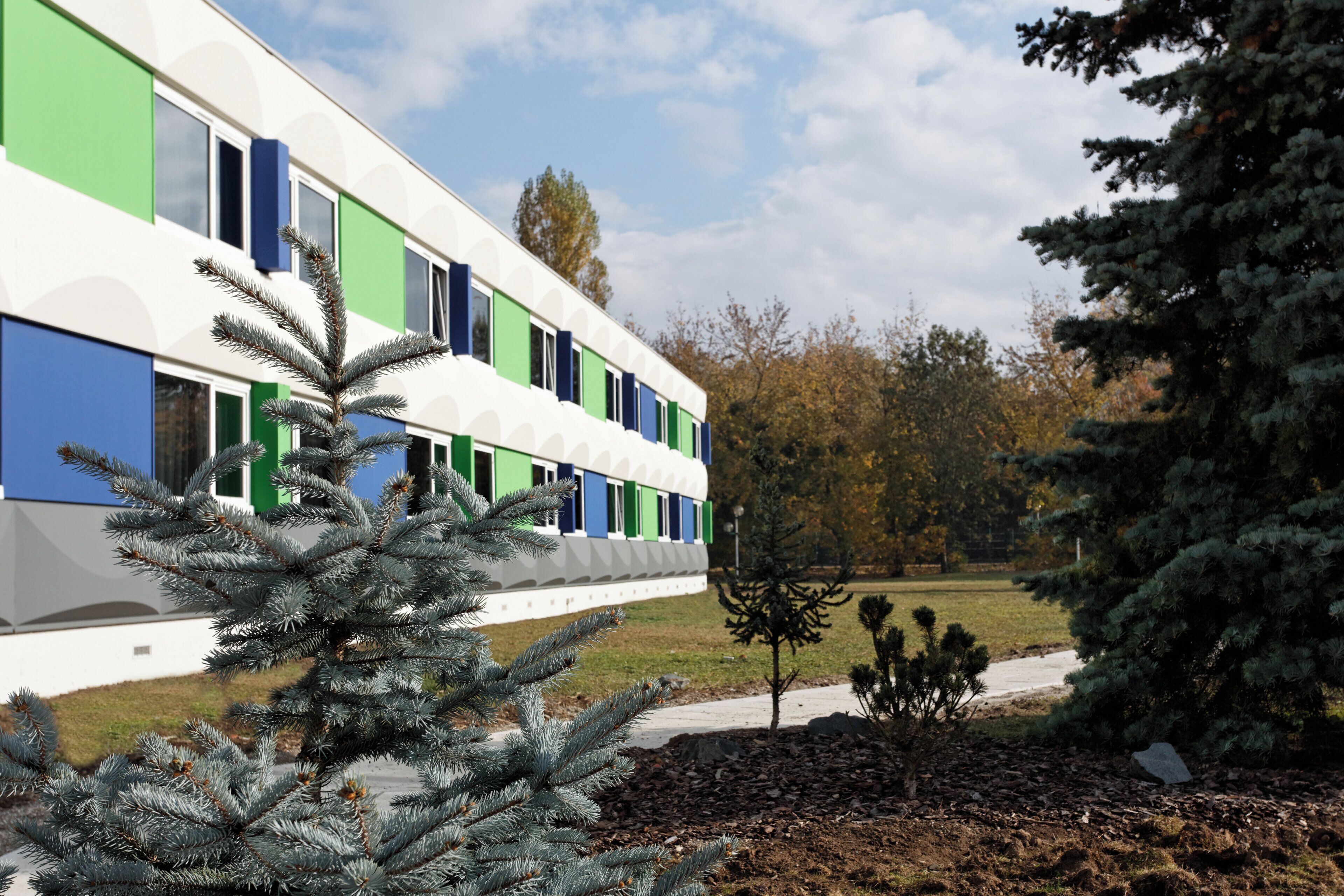 A modern building with a white facade and green and blue accents, surrounded by trees and a clear sky.