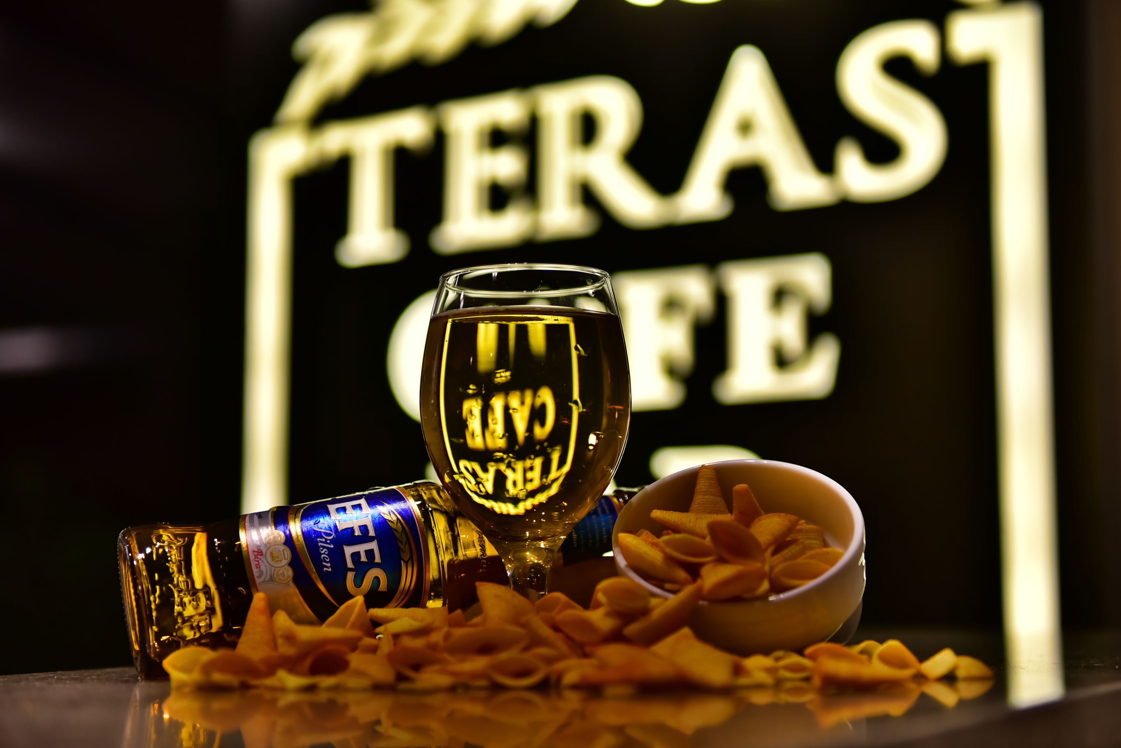 A glass of beer, a bottle, and a bowl of snacks on a reflective surface with a neon sign in the background.