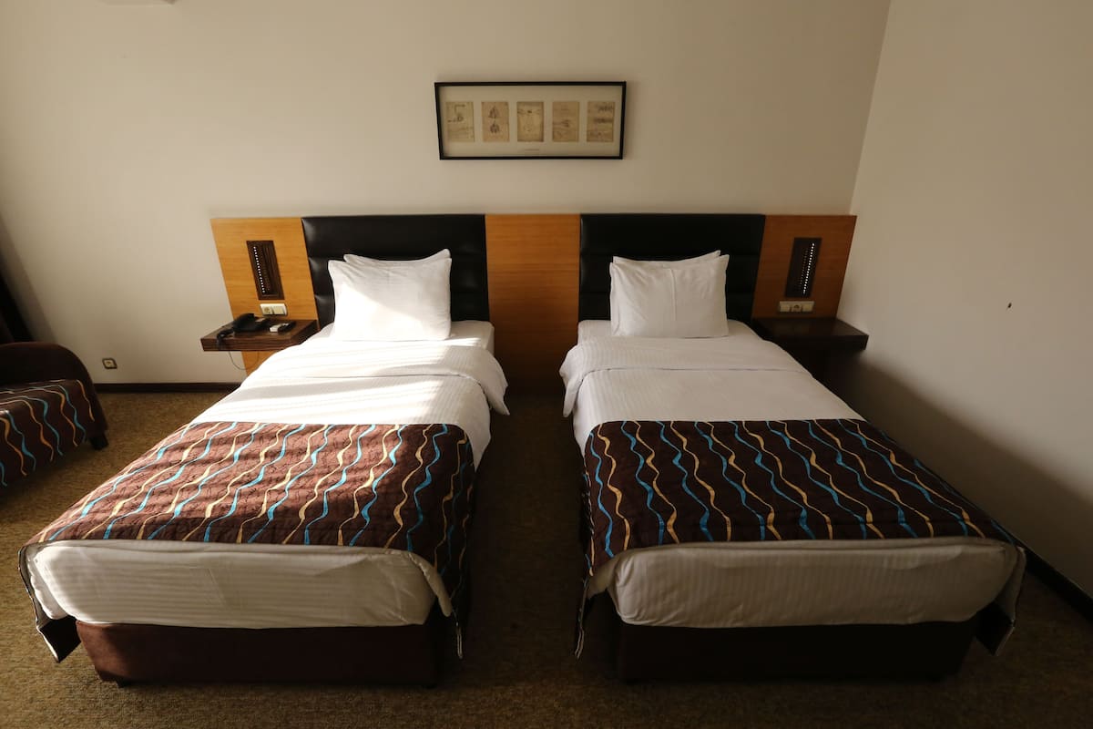 A hotel room with two single beds, a wooden headboard, a framed picture on the wall, and a nightstand with a phone and a lamp.