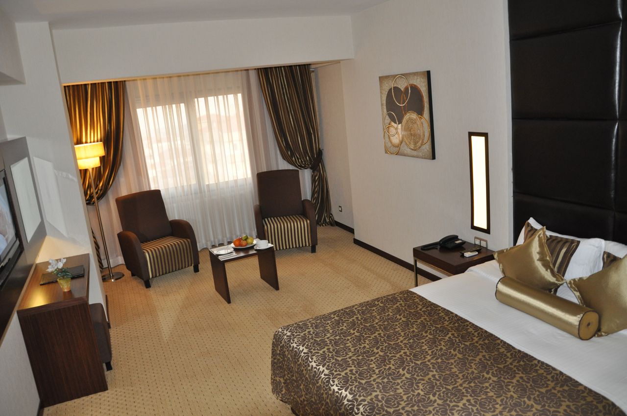 Een hotelkamer met een bed, twee fauteuils, een kleintje tafeltje met een schaal fruit, een bureau met een lamp, en een schilderij aan de muur.