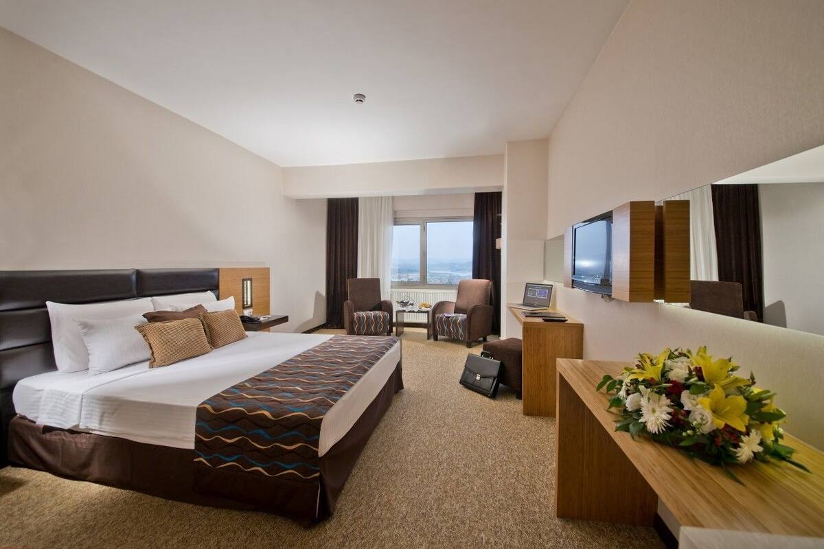A hotel room with a large bed, two armchairs, a desk with a computer, a TV, and a view of the city.