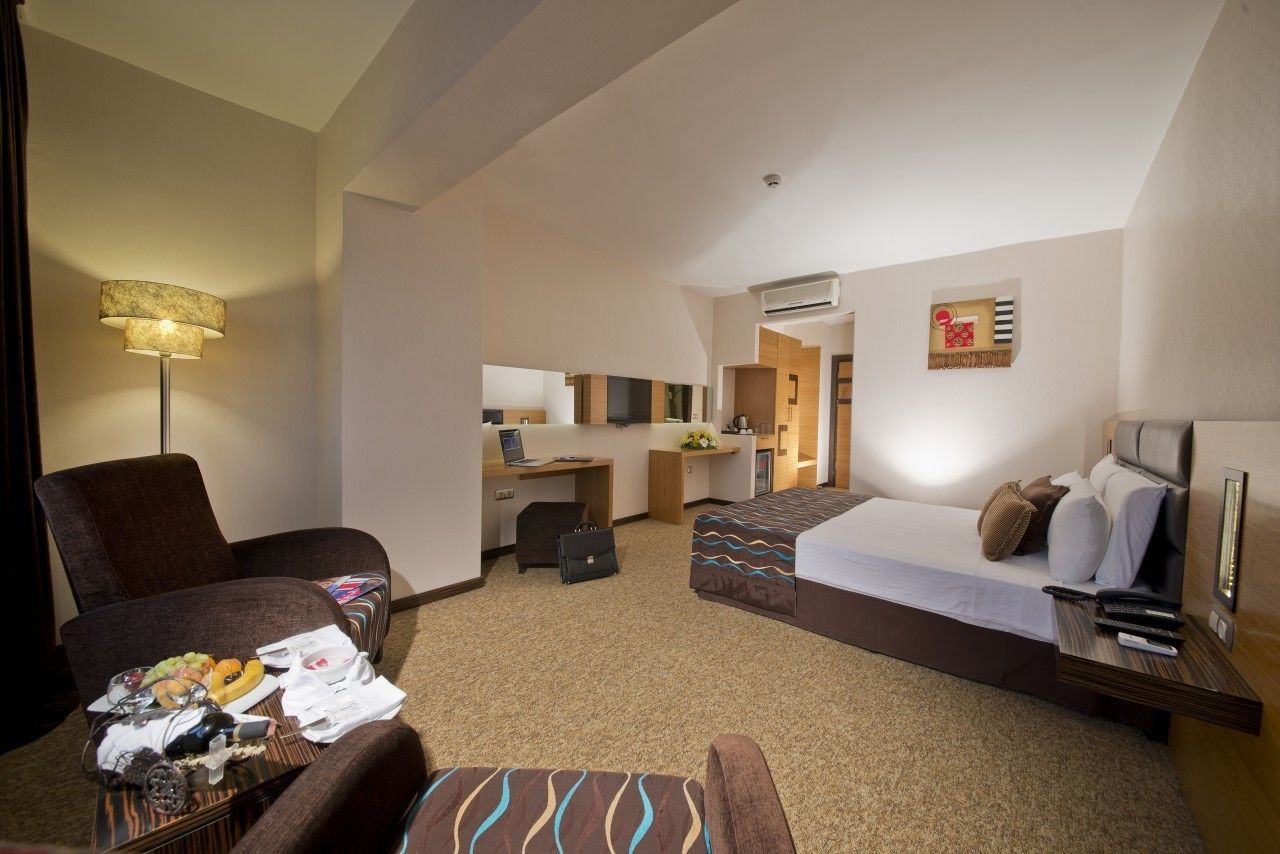 A hotel room with a bed, two armchairs, a desk with a computer, a small kitchenette, and a bathroom.