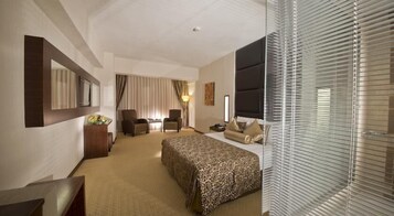 Habitación de hotel con una cama grande, dos sillones, un escritorio, un espejo y una ventana con persianas.