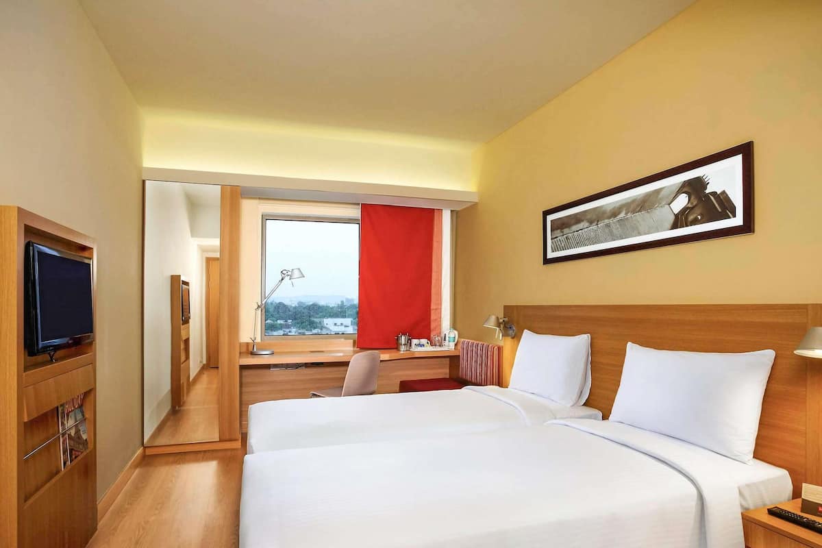 Ein Hotelzimmer mit zwei Betten, einem Schreibtisch, einem Fernseher und einem Fenster mit rotem Vorhang.