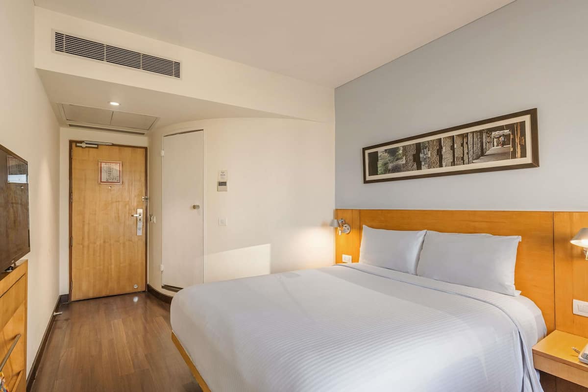 Ein Hotelzimmer mit einem Bett, einem Holzkopfstück, einem Nachttisch, einer Wandlampe, einer Tür mit Griff, einem Lüftungsgitter und einem gerahmten Bild an der Wand.