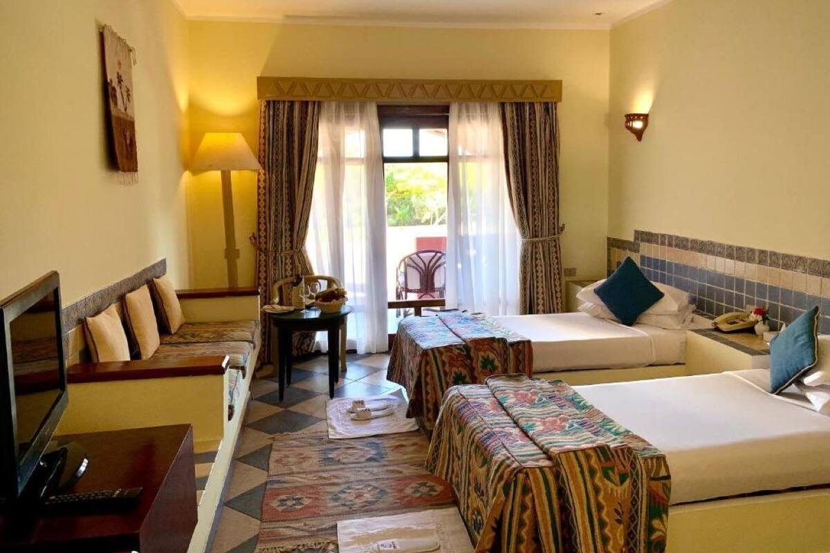 Habitación de hotel con cama, sofá, escritorio y una silla. Se ve una ventana con cortinas, una lámpara y un aplique de pared.