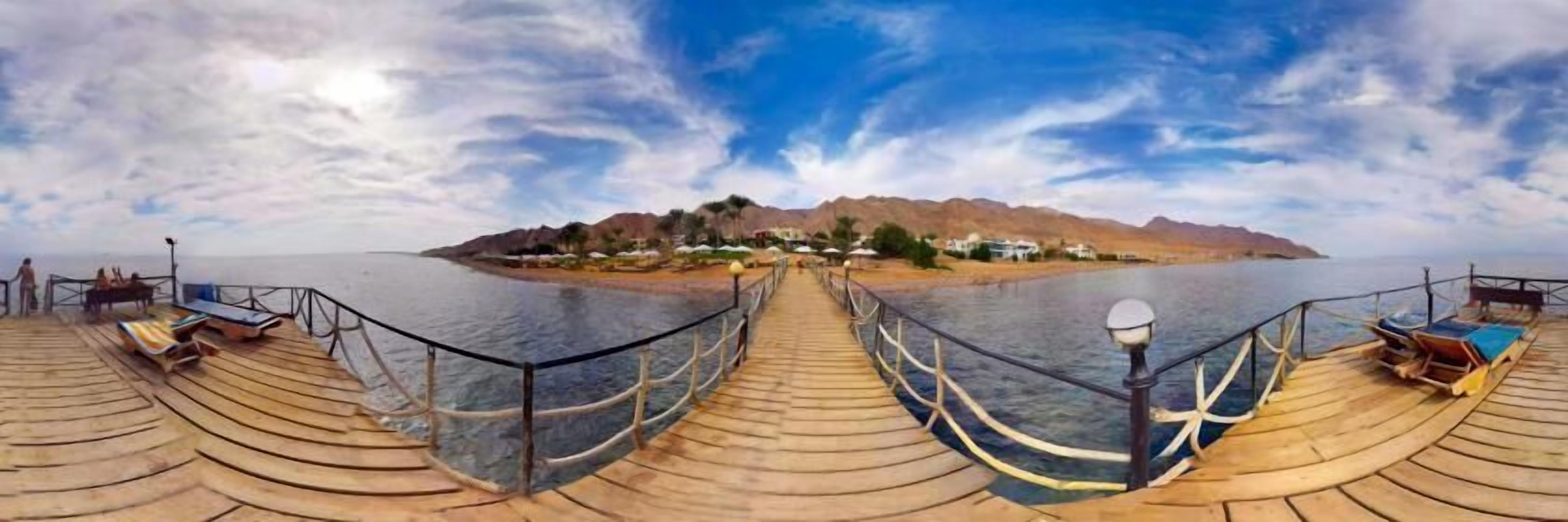 Une terrasse en bois, dotée de rampes de protection, mène à un plan d’eau et offre une vue sur une petite île où se trouvent des bâtiments et des palmiers.