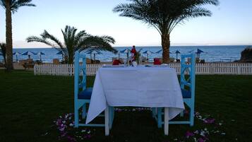 An outdoor dining setup with a white table and blue chairs, surrounded by palm trees and a view of the sea.