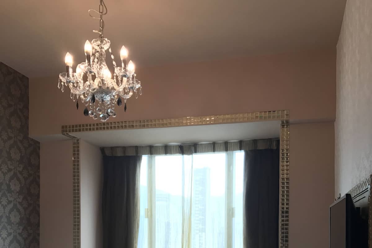 Une chambre d’hôtel avec un grand lit, un lustre, une fenêtre avec des rideaux, une télévision et un bureau équipé d’un téléphone et d’une machine à café.