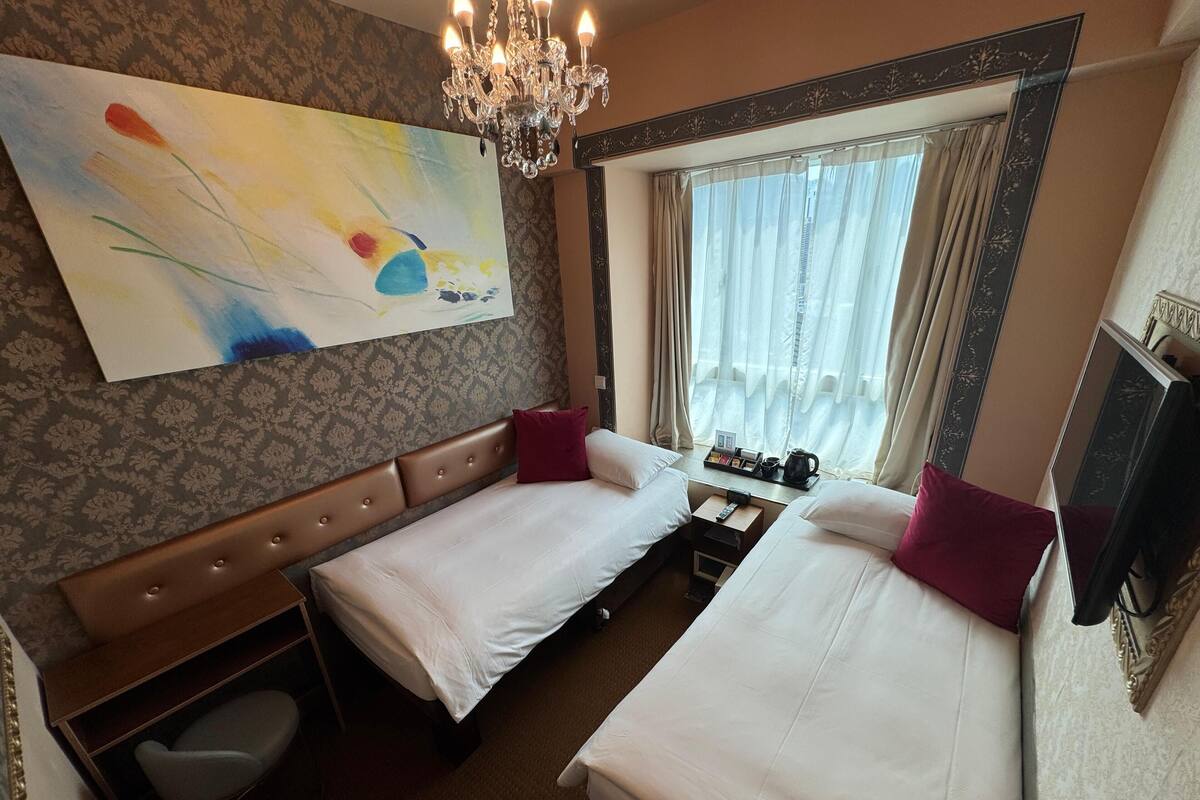 Une chambre d’hôtel avec deux lits, un tableau, un lustre, une fenêtre avec des rideaux et une petite table avec une télécommande.