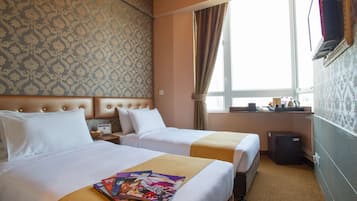 Quarto de hotel com duas camas, uma pequena escrivaninha, televisão e janela com cortinas.