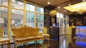 Un hall d’entrée luxueux, avec un canapé doré et des fauteuils richement décorés, de grandes fenêtres, un comptoir d’accueil et un lustre.