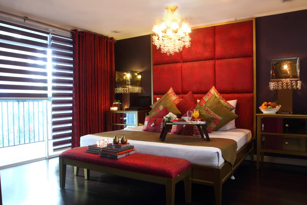 A hotel room with a large bed, a bedside table with a lamp, a bench, and a view of the outdoors through large windows.