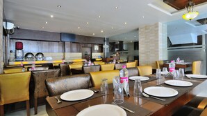 Un restaurant au décor moderne, avec une longue table à manger dressée, où l’on trouve des assiettes, des couverts et des bouteilles d’eau.