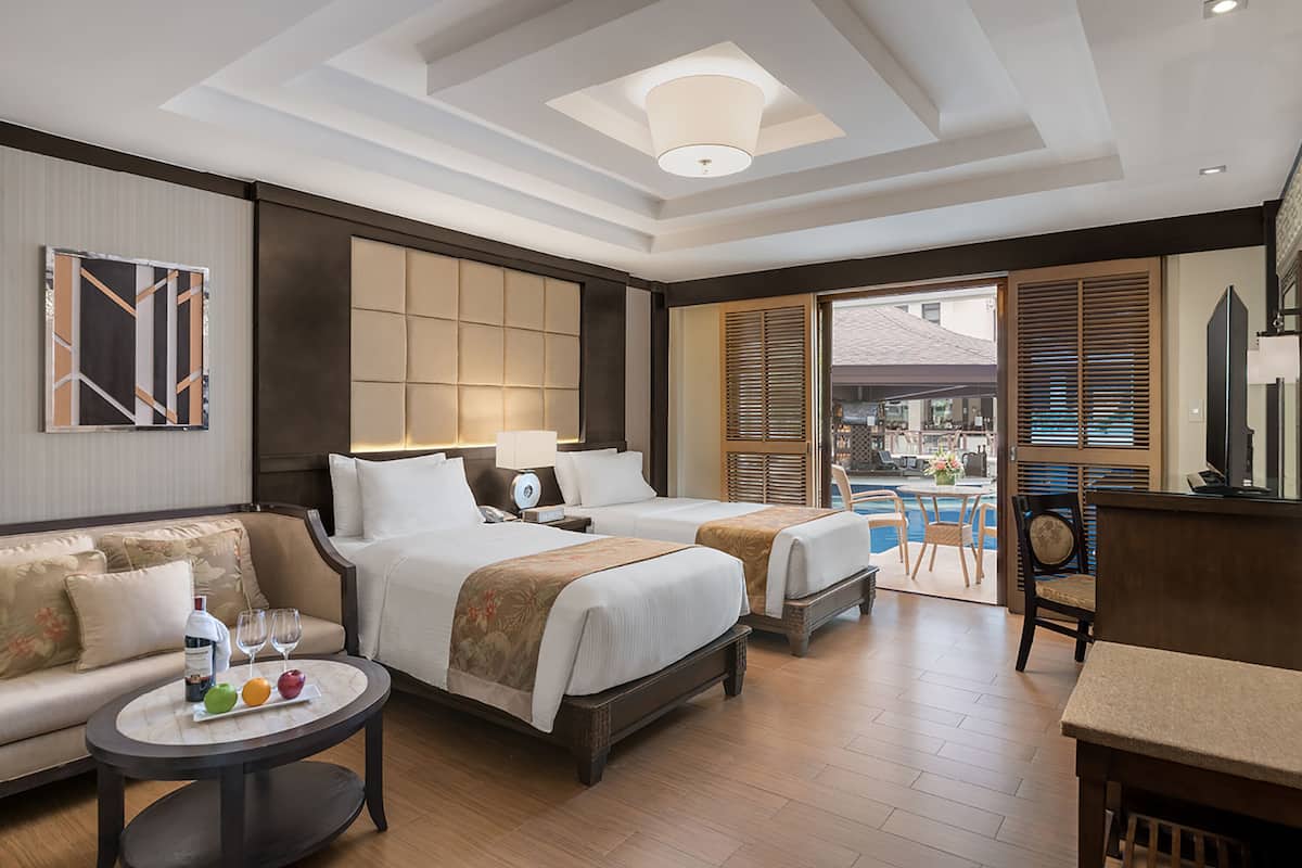 A hotel room with two beds, a sofa, a small table with fruit and a bottle, and a view of the outside.