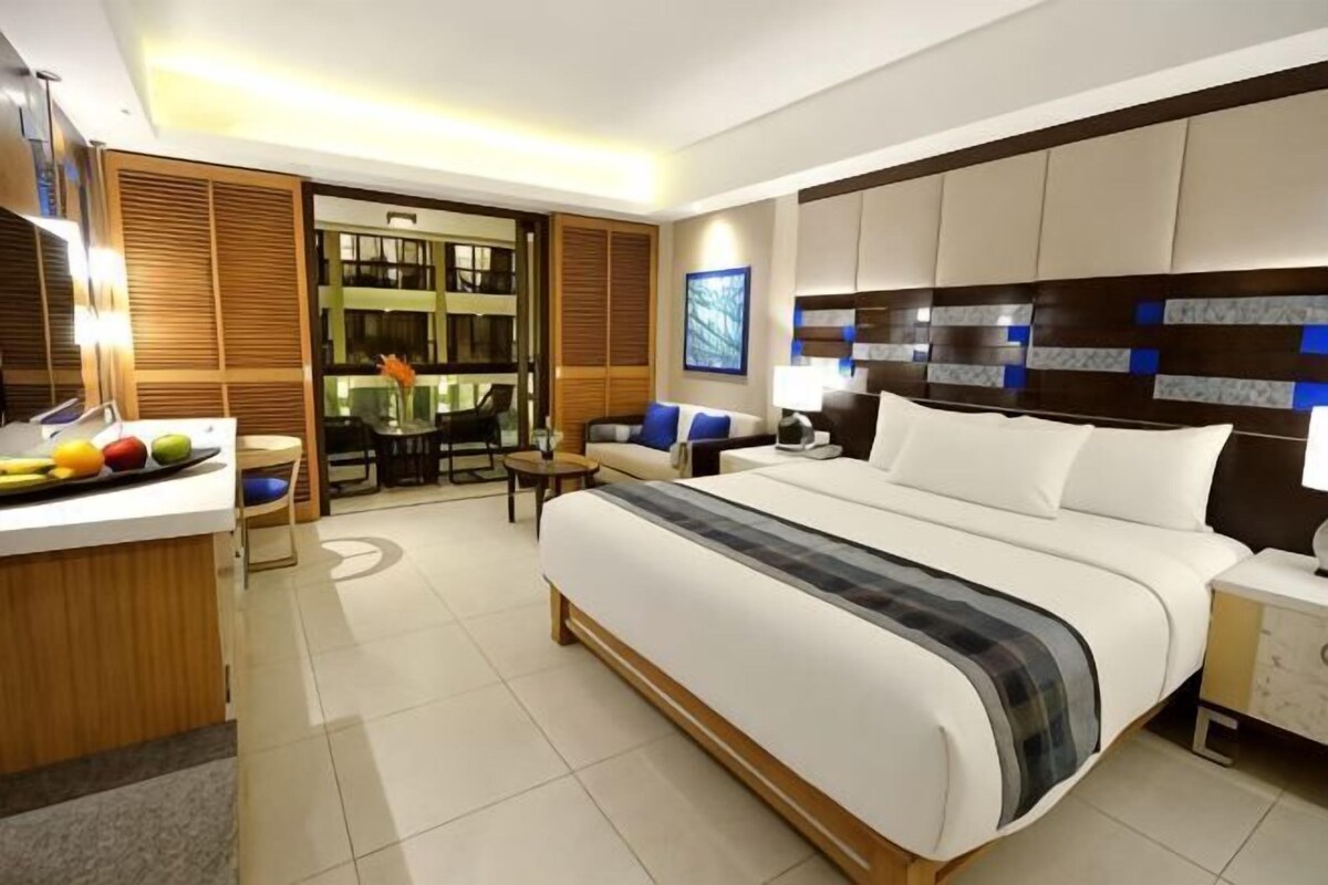 A modern hotel room with a large bed, a bedside table, a desk with a chair, and a wardrobe.