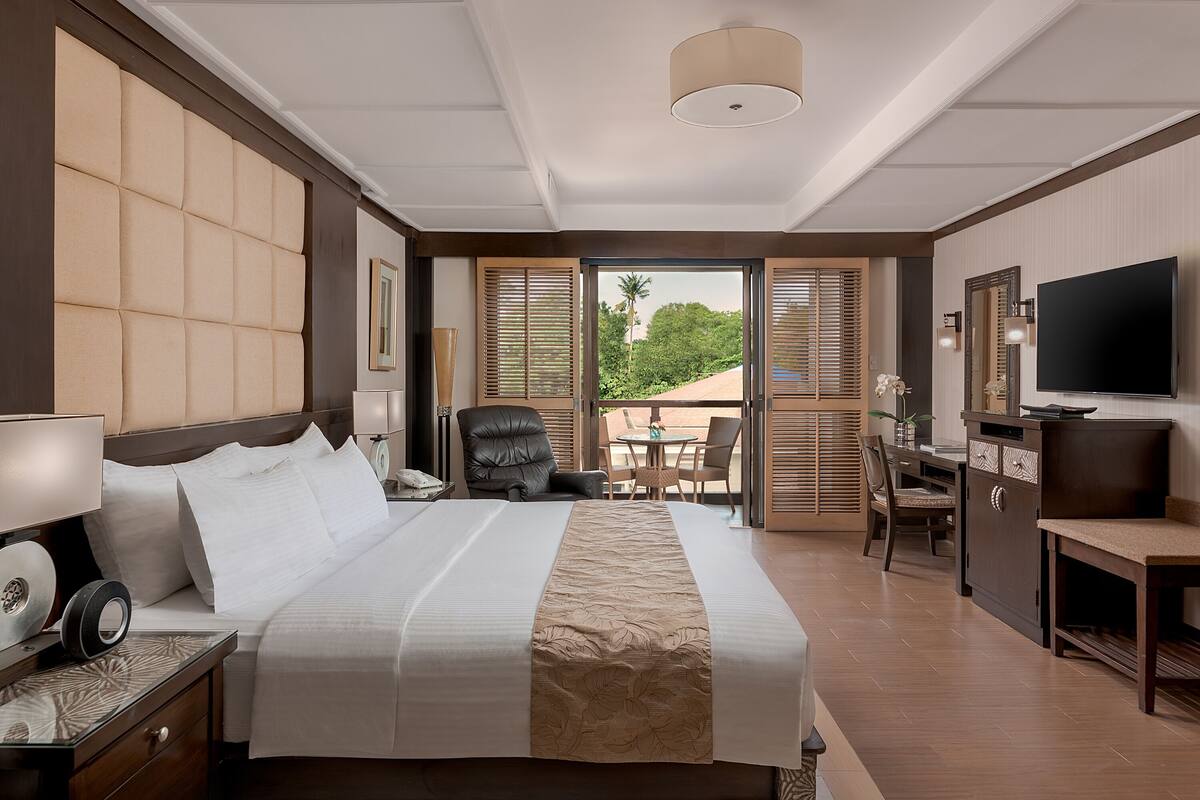 A hotel room with a large bed, a desk, a chair, a TV, and a balcony with a view of trees.