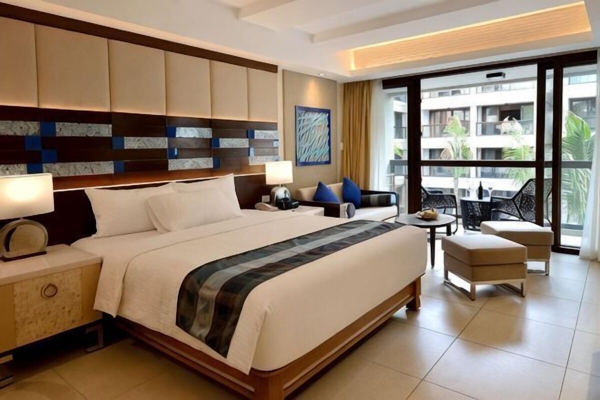 A hotel room with a large bed, bedside tables, a small table, a chair, and a balcony with a view.