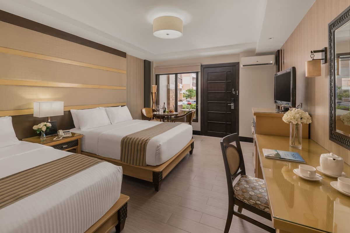 A hotel room with two beds, a desk with a chair, a TV, and a window with a view of buildings.