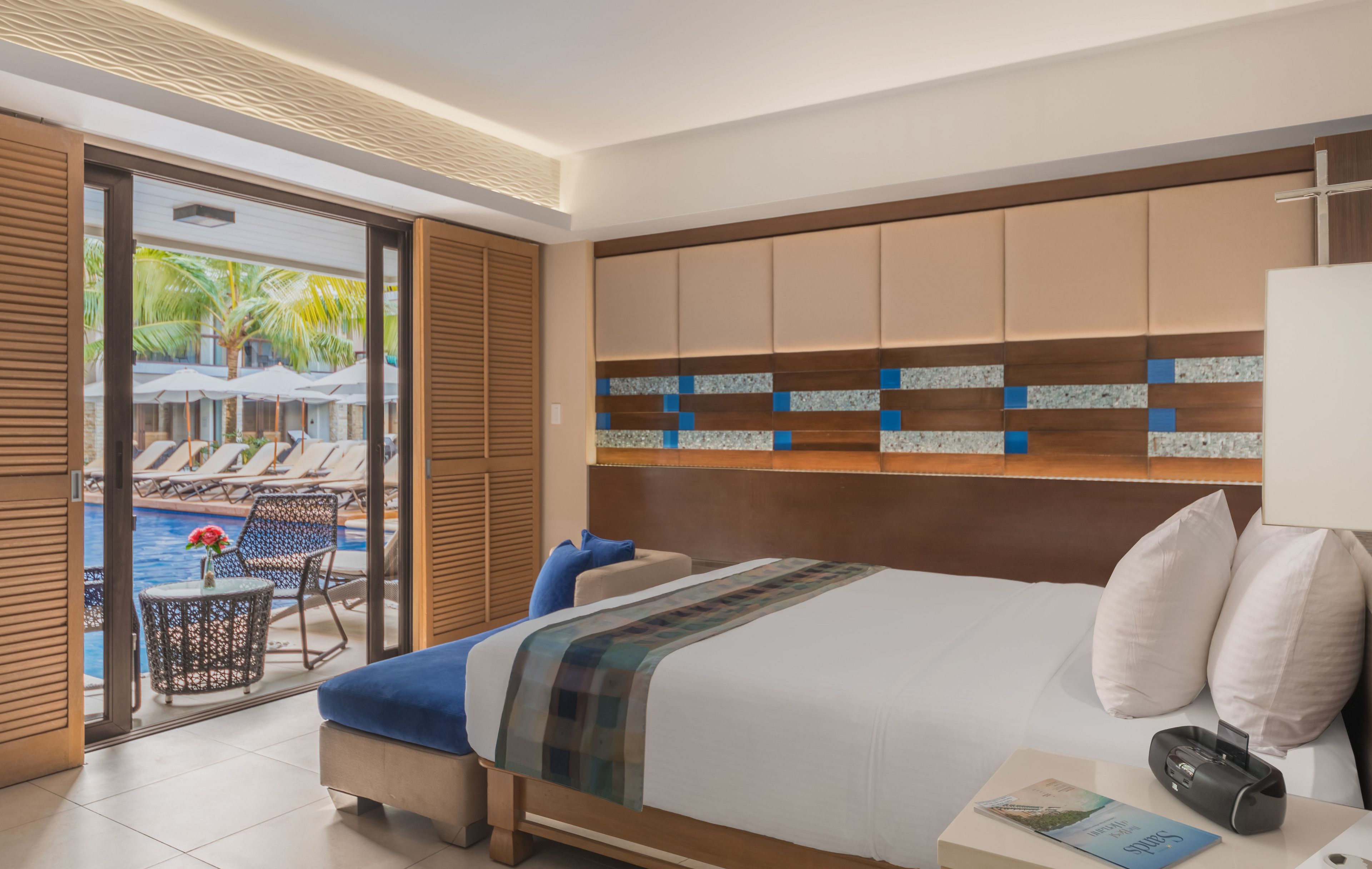Quarto de hotel com uma cama grande, mesa de cabeceira, abajur e vista para área da piscina com espreguiçadeiras e guarda-sóis.