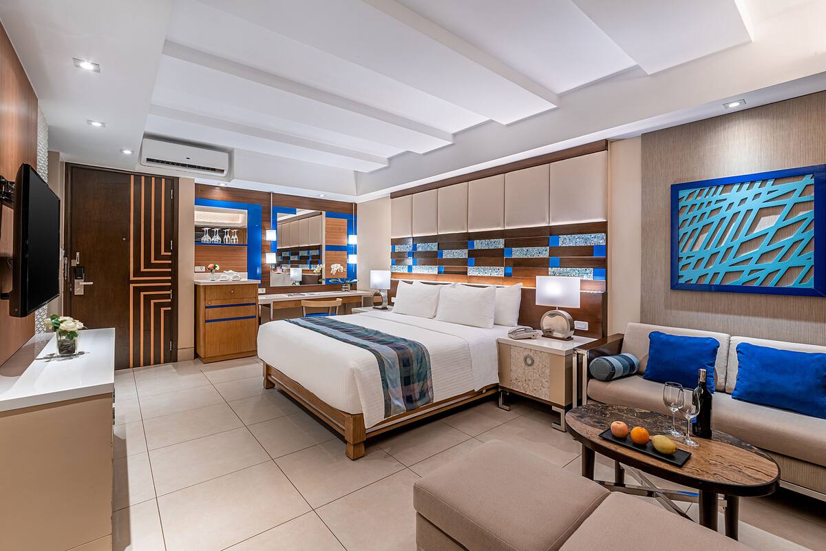 A modern hotel room with a large bed, a sofa, a coffee table with fruits and a bottle, a desk with a computer, and a bathroom with a mirror and shelves.