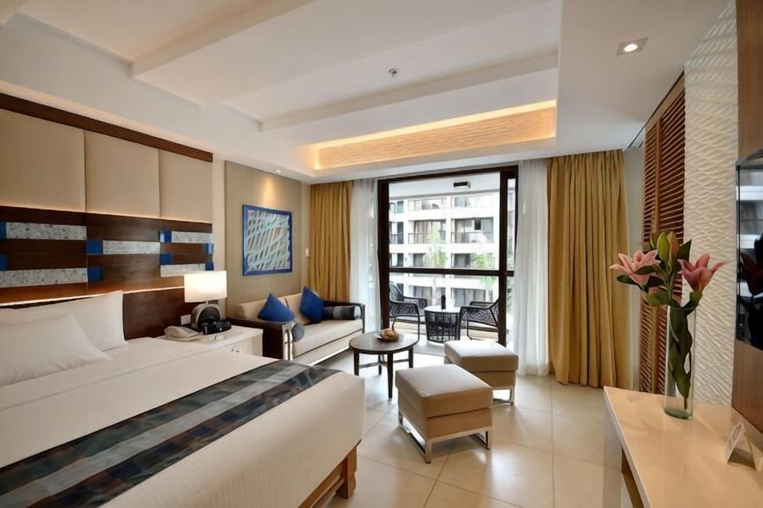 A hotel room with a bed, a sofa, a small table, a chair, a desk, a lamp, a vase with flowers, and a large window with a view of buildings.