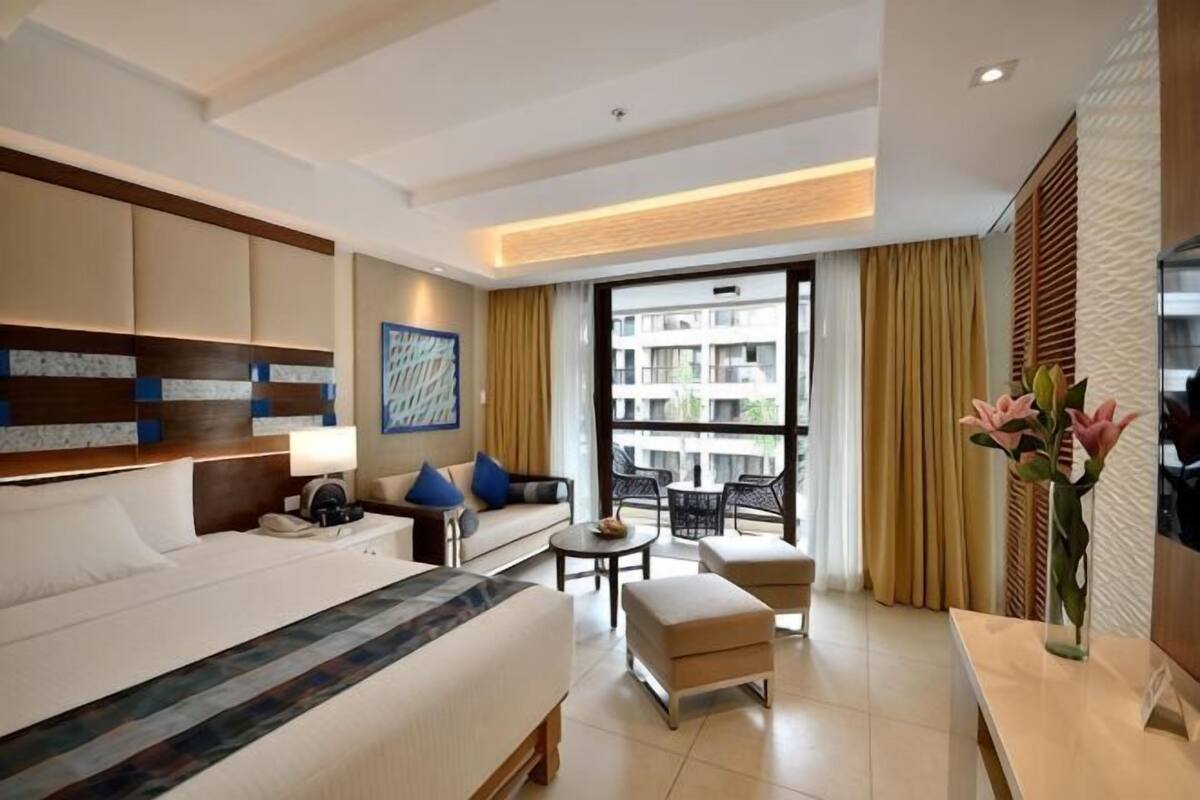 A hotel room with a bed, a sofa, a small table, a chair, a desk, a lamp, a vase with flowers, and a large window with a view of buildings.
