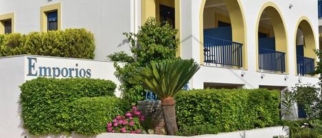 A building with a sign reading 'Emporios,' featuring a white facade with yellow arches, a palm tree, and pink flowers.