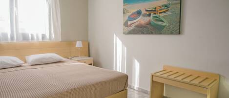 A neatly made bed with a patterned bedspread, a bedside table with a lamp, a wooden bench, and a beach-themed painting on the wall.