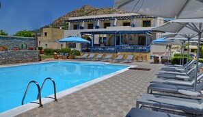 Une piscine avec des chaises longues et des parasols, un mur en pierre et un bâtiment aux accents bleus.
