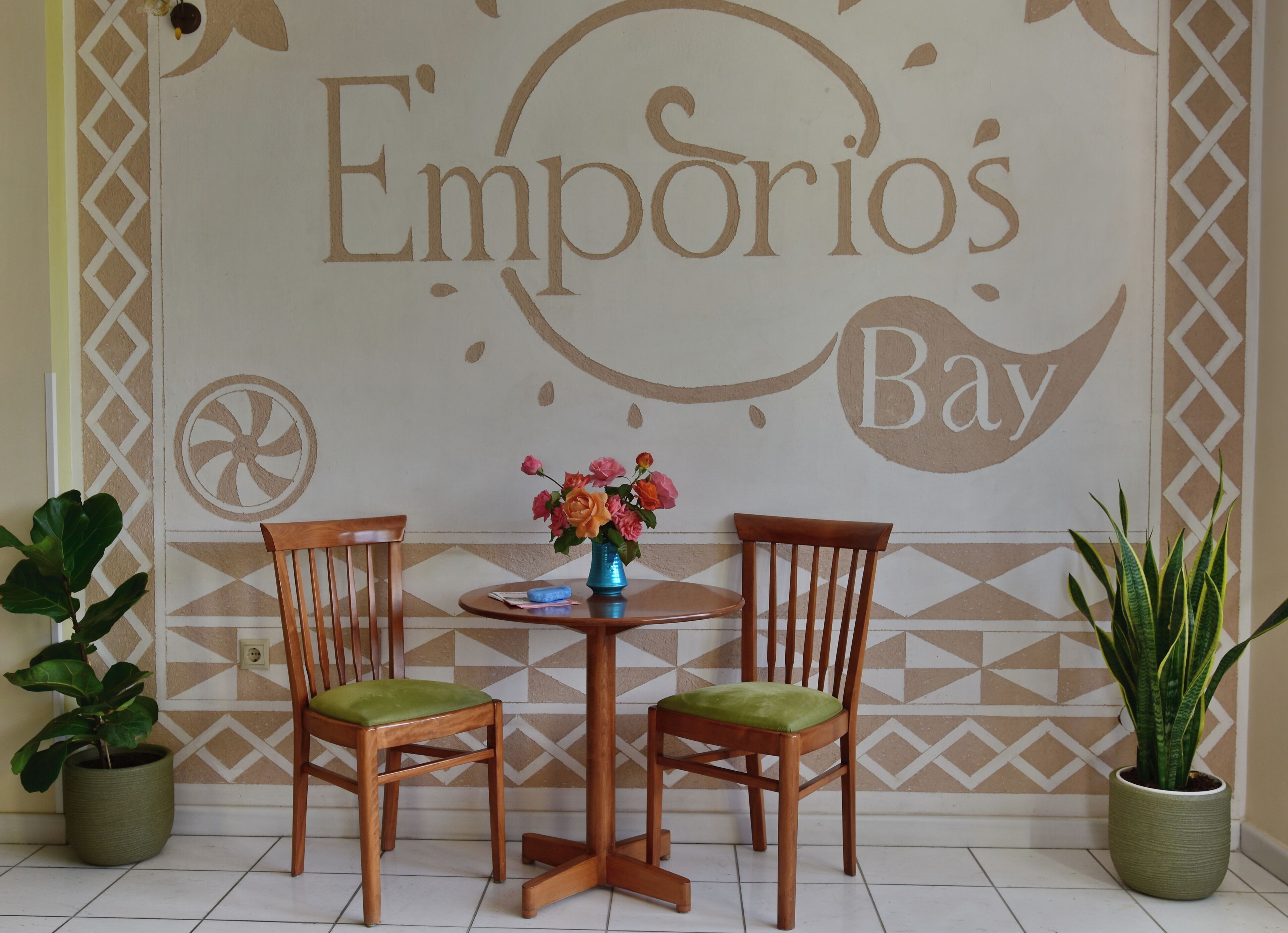 A room with two wooden chairs, a small round table, and a vase with flowers. The wall has a geometric pattern and the words 'Empório's Bay' are visible.