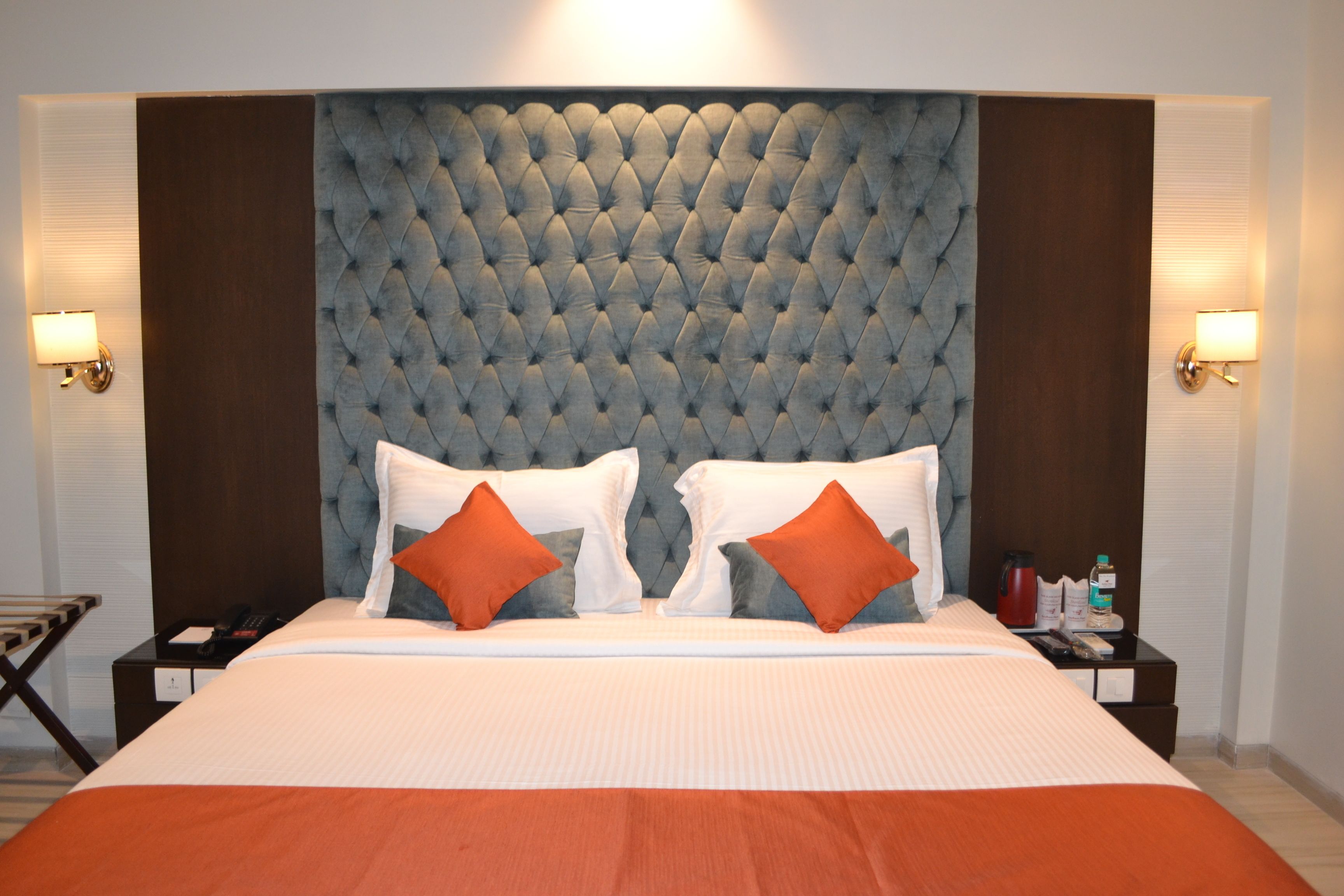 A hotel room with a tufted headboard, a large bed with white and orange bedding, two bedside tables with lamps, and a nightstand with a telephone and a bottle.