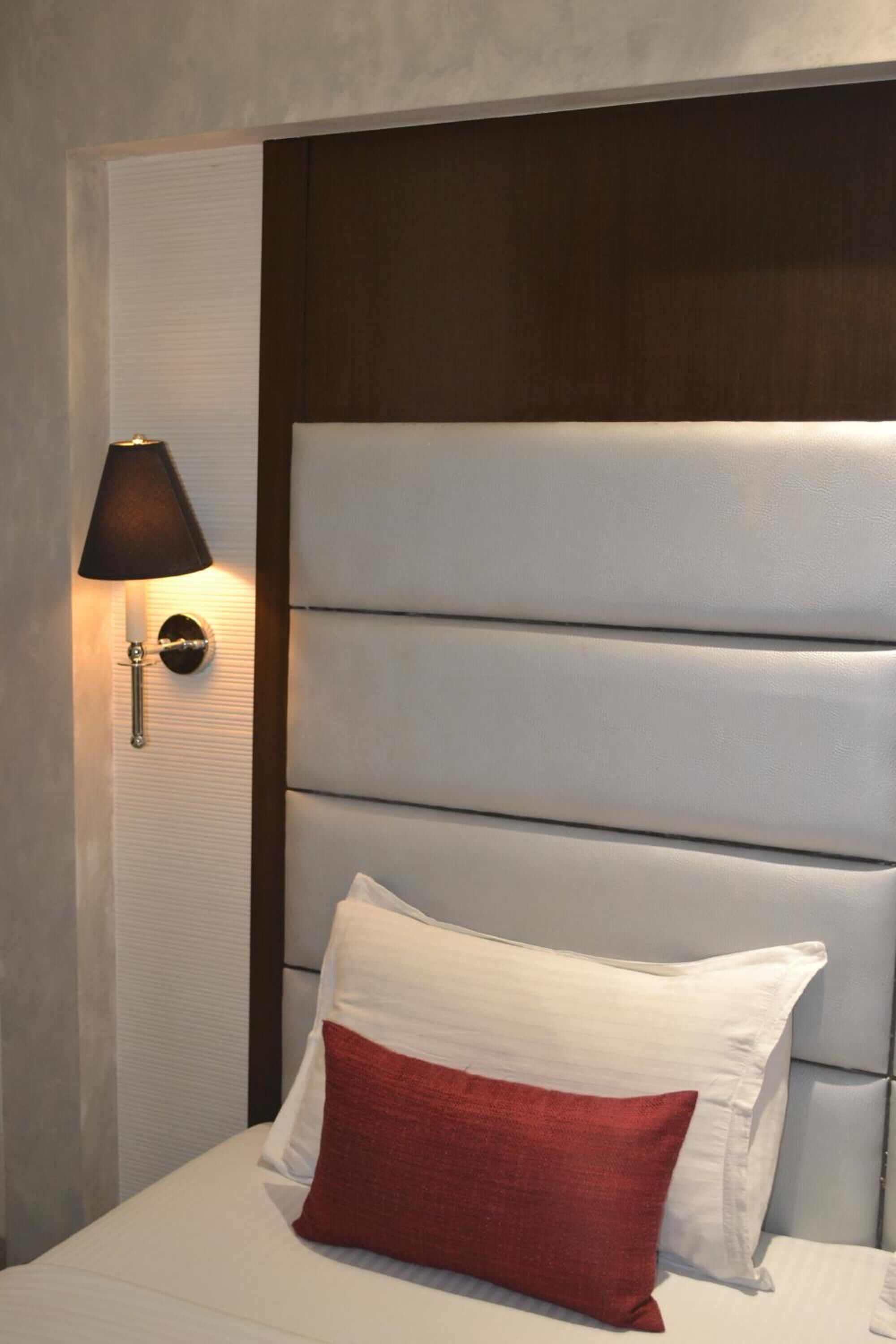 A hotel room with a padded headboard, a bedside lamp, and a bed with white and red pillows.