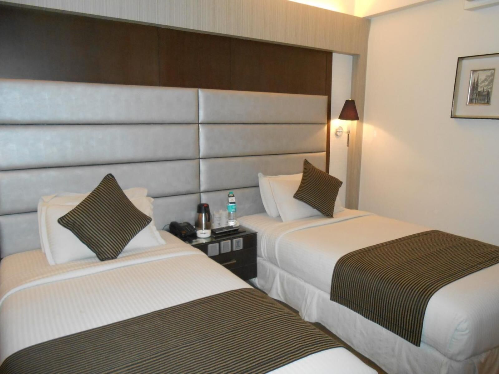 Ein Hotelzimmer mit zwei Betten, jeweils mit Kopfteil, Nachttisch mit Lampe und einem gerahmten Bild an der Wand.
