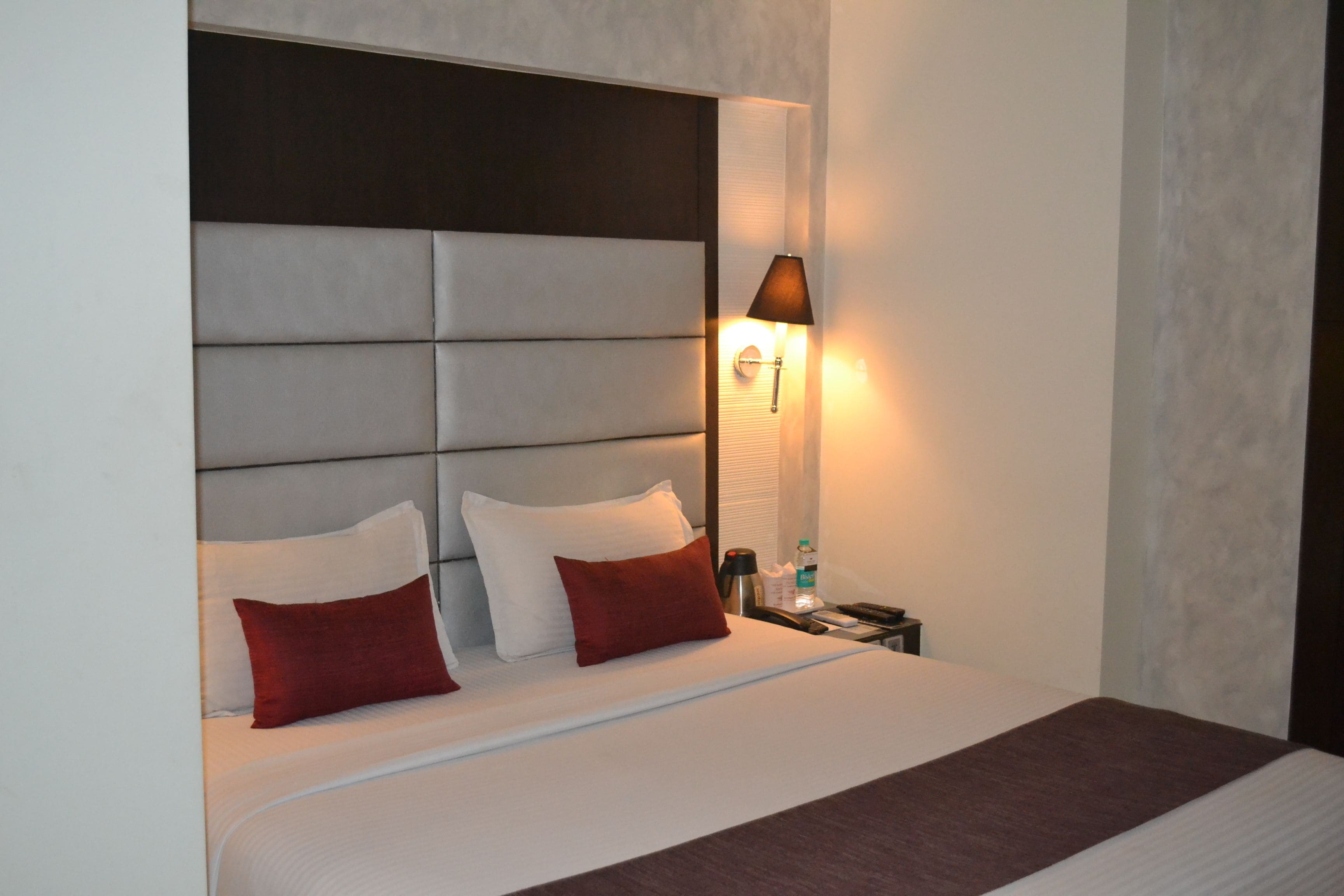 A neatly made bed with a headboard, two pillows, a bedside table with a lamp, and a bottle of water.