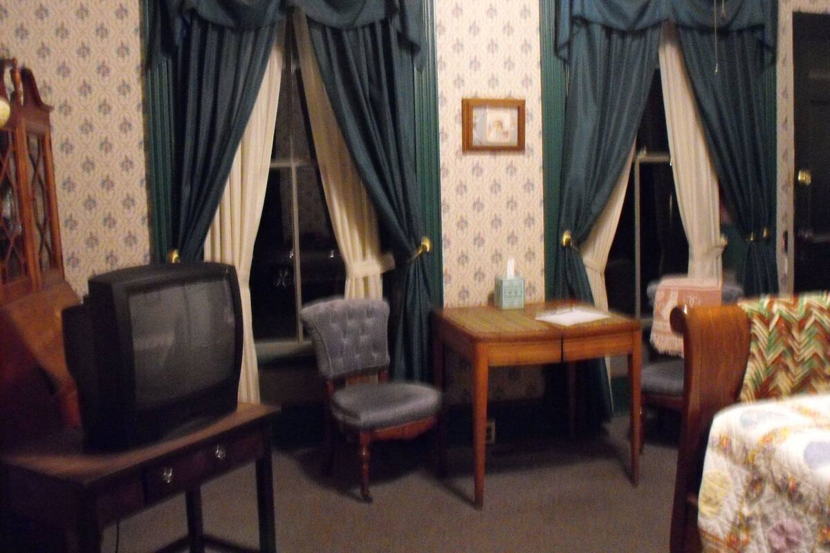 Ein kleiner, schwach beleuchteter Raum mit einem Fernseher, zwei Stühlen, einem kleinen Tisch und einem Bett.