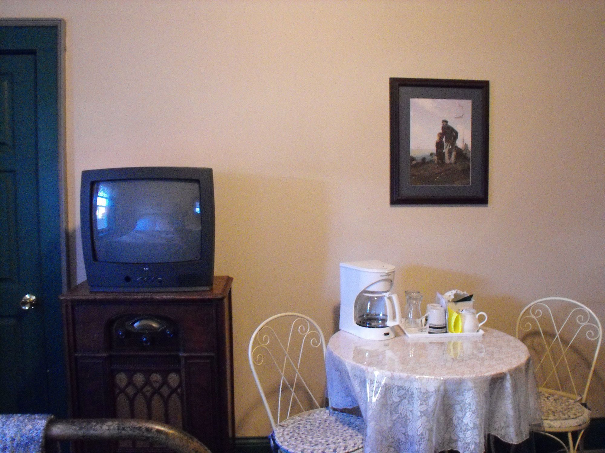 Una habitación con televisor sobre un soporte de madera, cafetera y una mesa con sillas.