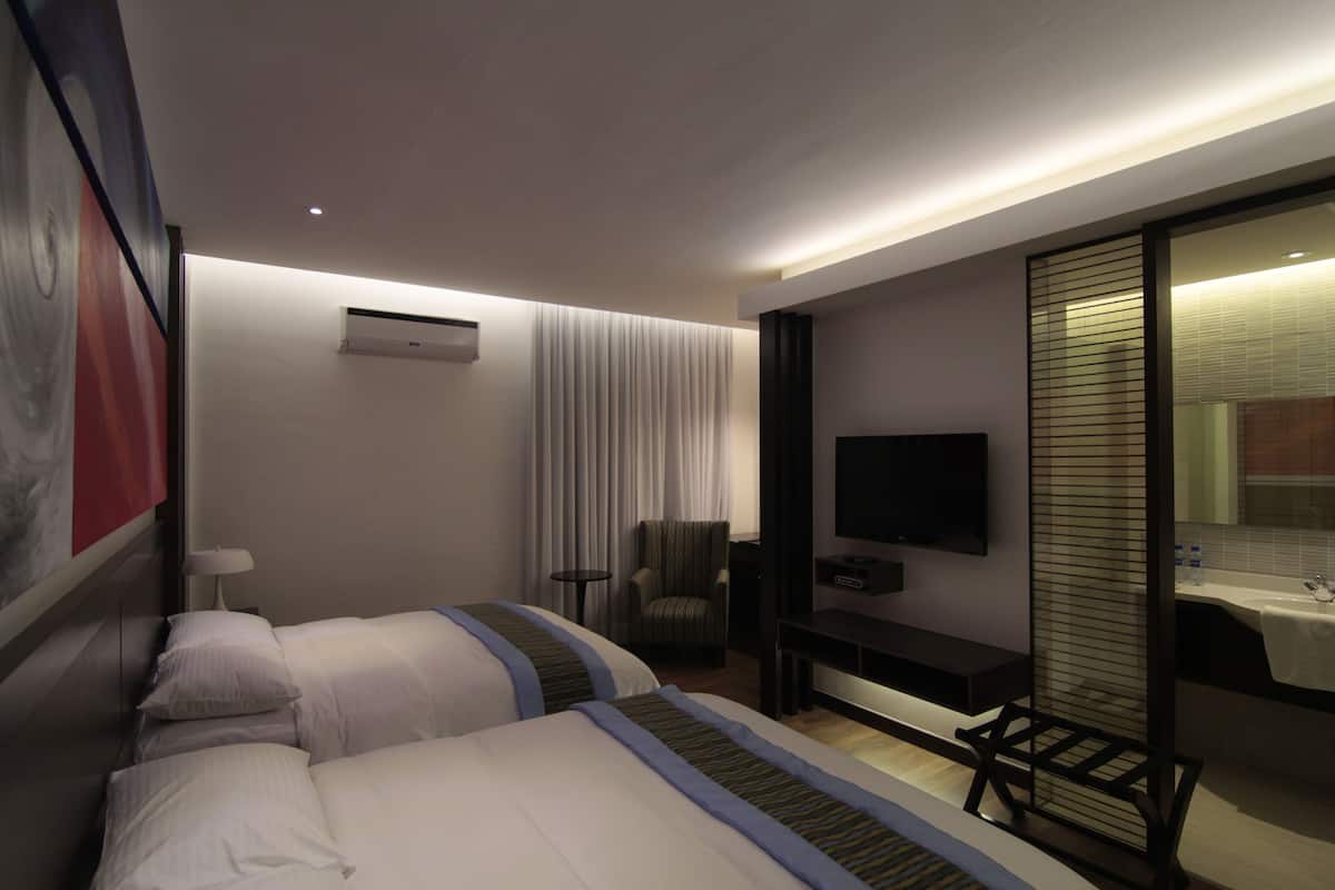 A hotel room with two beds, a desk, a chair, a TV, and a bathroom with a sink and mirror.