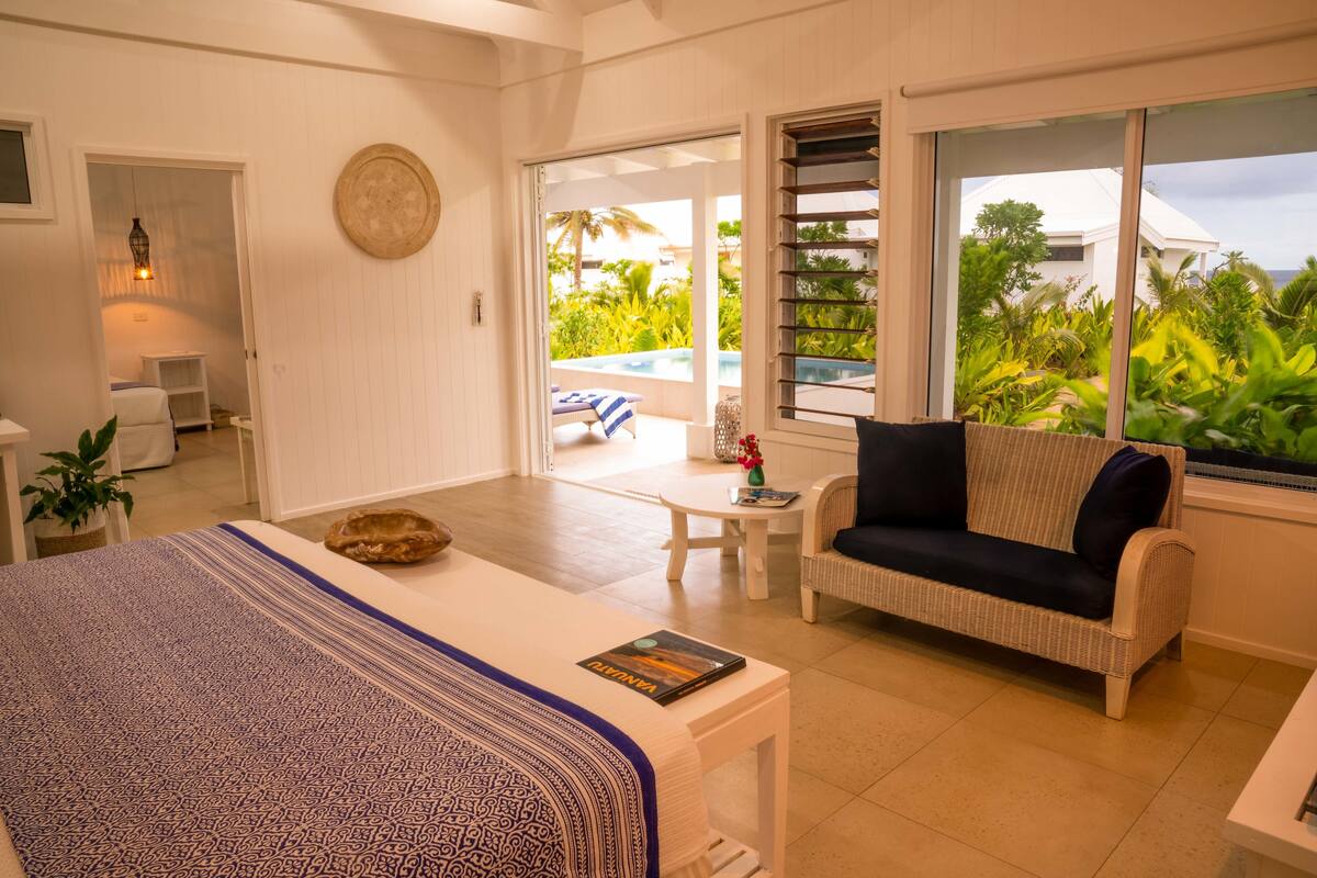 A spacious room with a bed, a wicker sofa, a small table, and a view of a pool and greenery outside.